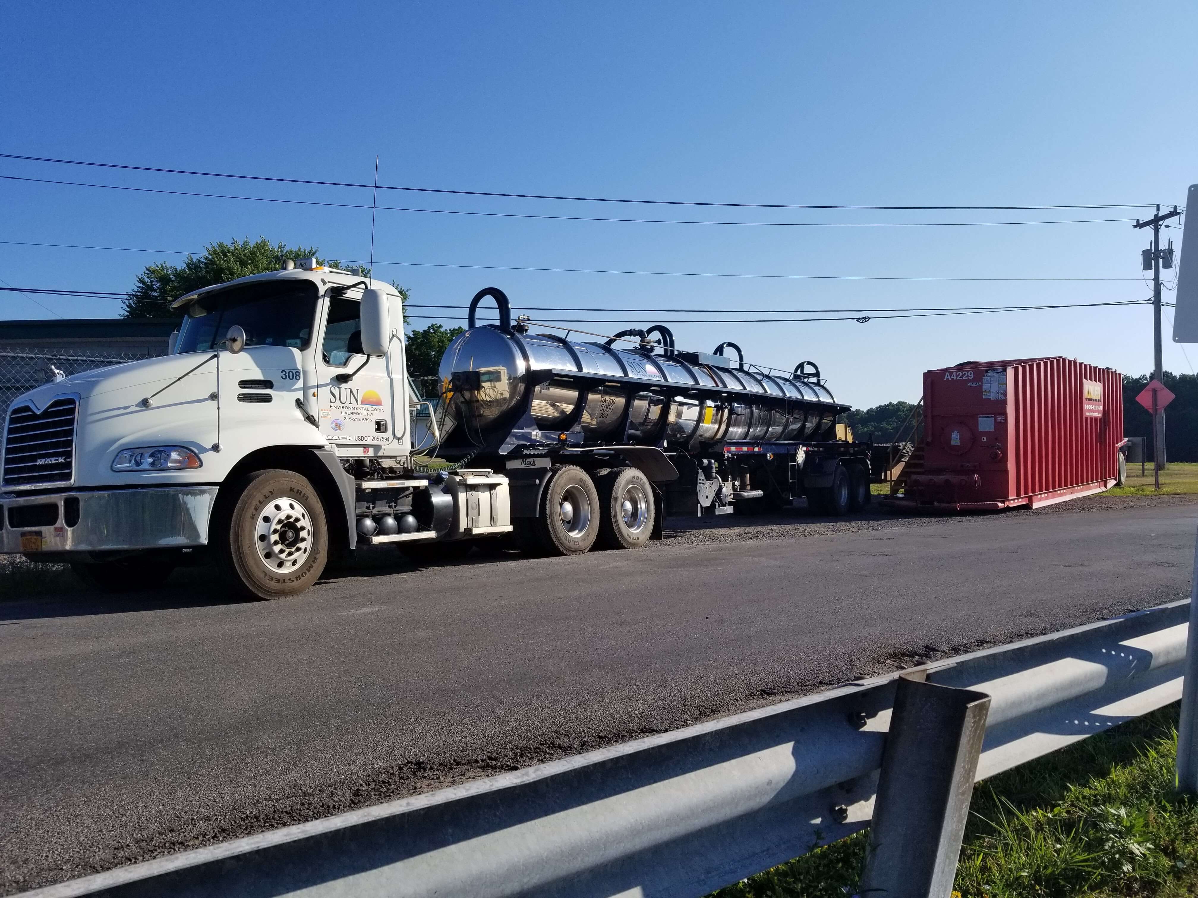 A white semi-truck with the Sun Environmental Corp. logo on the door and long, silver tank on the back is parked next to a red hazardous waste bin as it vacuums up the liquids held inside the bin so it can safely haul it away for proper disposal.