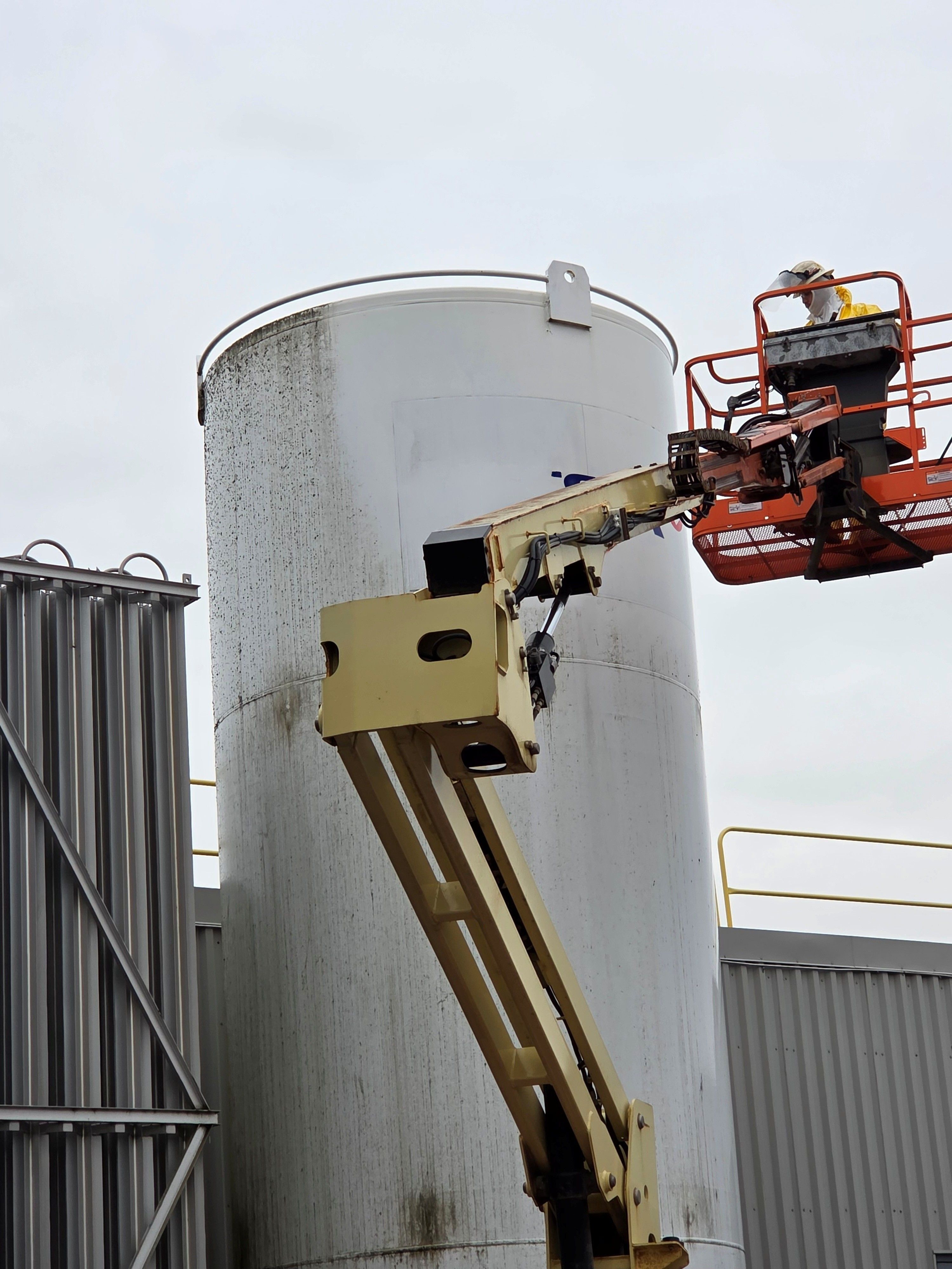 An employee of Sun Environmental Corp. is being raised in the air on a bucket truck in order to clean a tall white tank and they are about half way done with the job, so the part of the tank is looking nice and white and part is still looking very dirty.