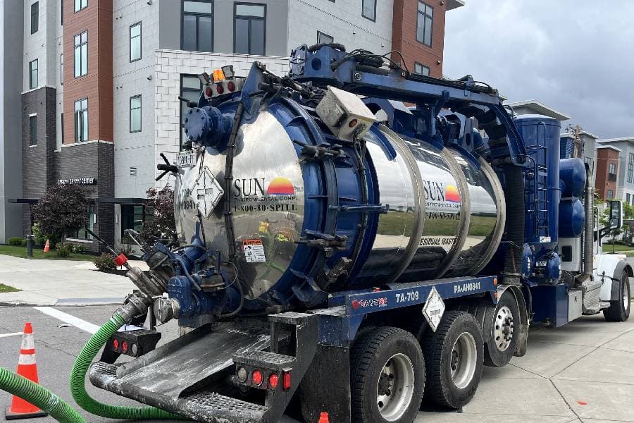 A large blue and silver vacuum truck with the Sun Environmental Corp logo on the side is parked on a street with a green hose coming out of the back that is used to vacuum up industrial or hazardous waste.